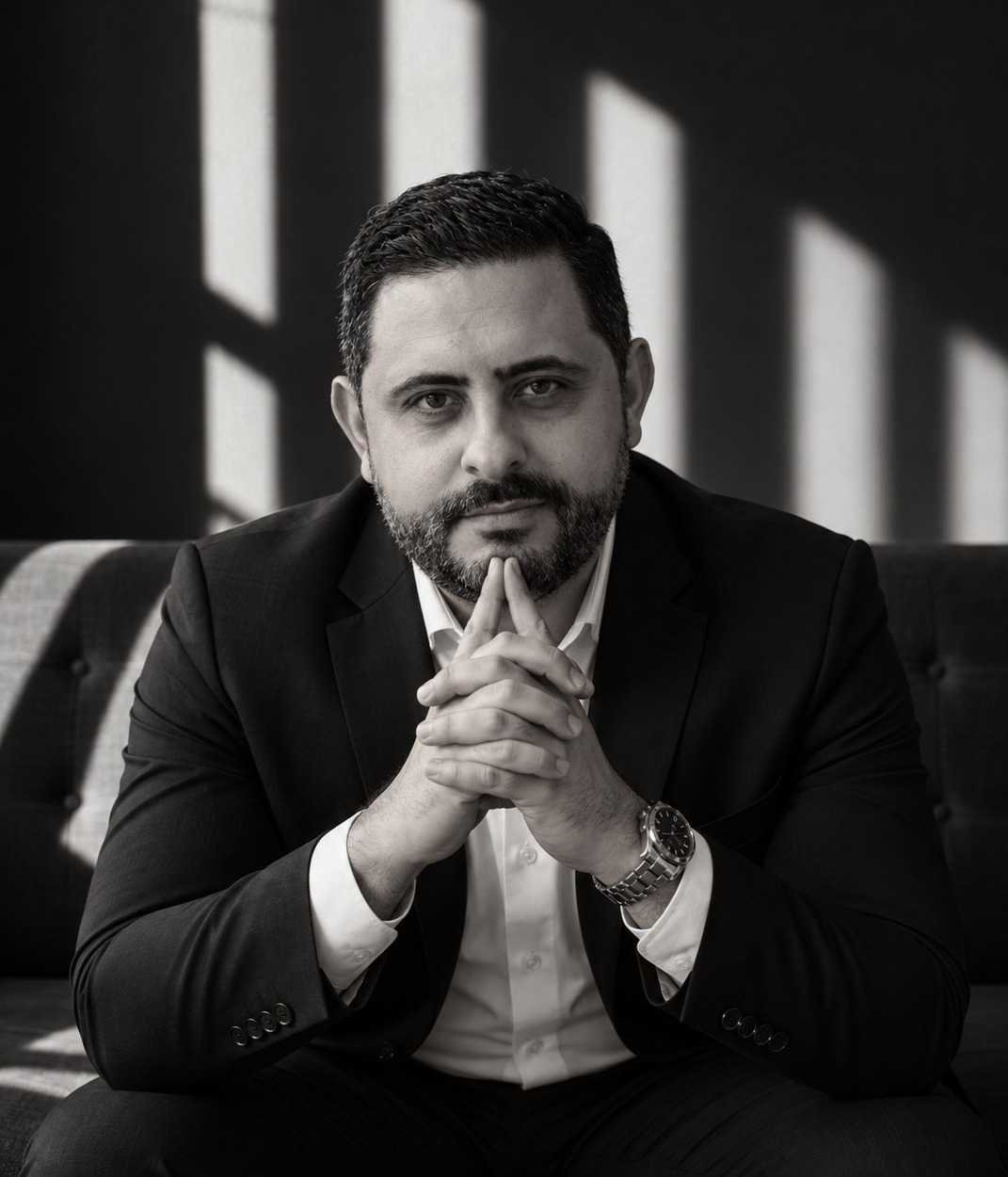 Isaac Mello em retrato preto e branco, sentado, com as mãos unidas, sob a luz de uma janela
