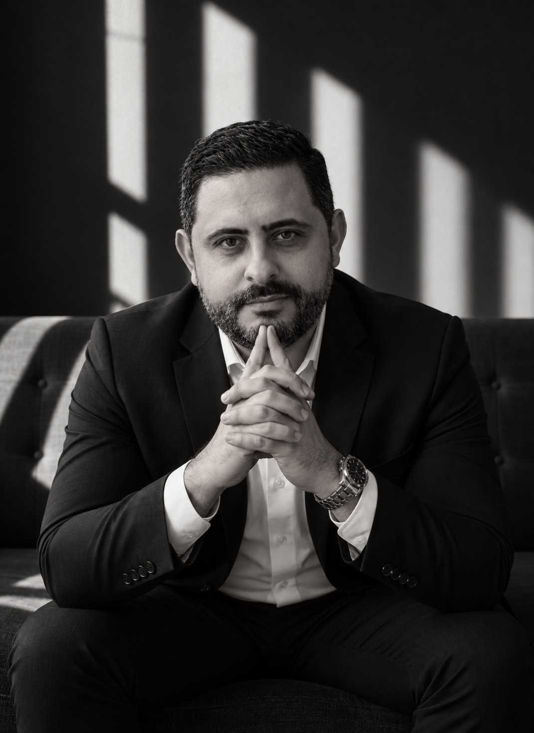 Isaac Mello, em retrato em preto e branco, sentado com as mãos unidas